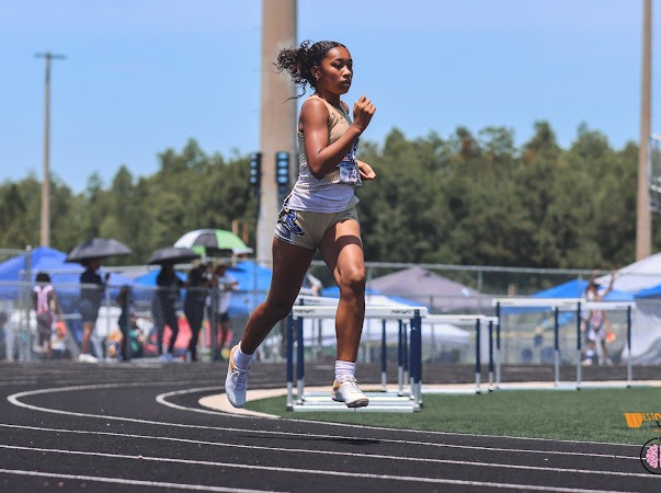 Athlete sprinting on a track during a competition, showcasing determination while hurdles and spectators are in the background.