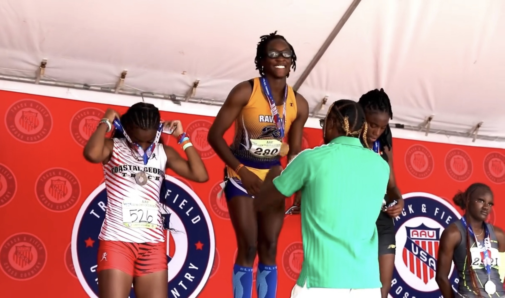Athletes celebrate on the awards podium at a track and field event, with one receiving a gold medal and two others preparing to wear their medals.