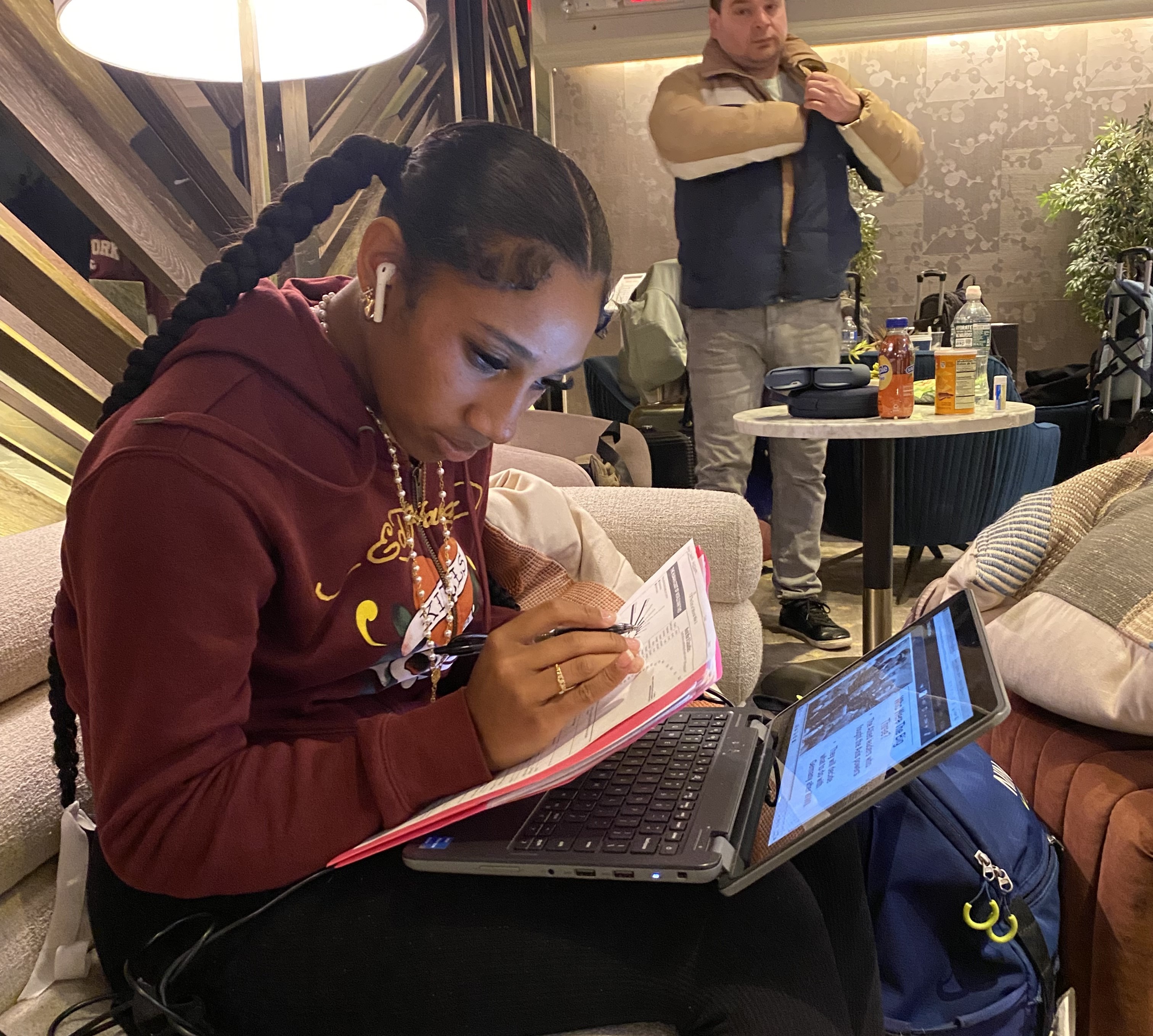 A focused individual with a long braid works on a laptop while taking notes on paper. A busy café setting is visible in the background.