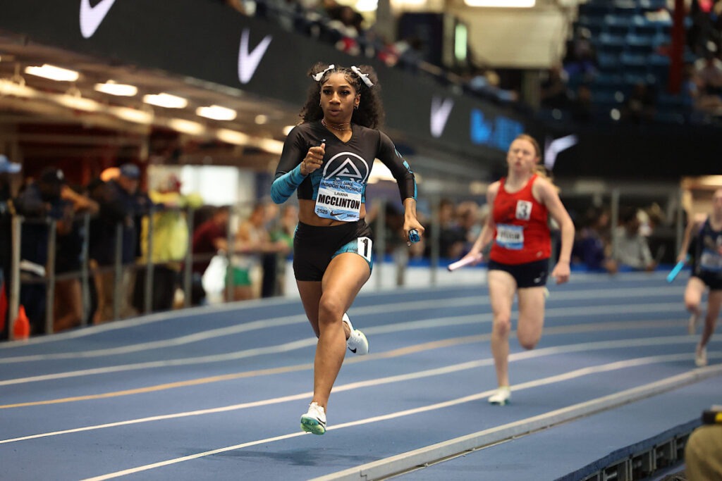 Female sprinter in a black and blue uniform races on a track, baton in hand, during a relay event. Spectators watch in the background.