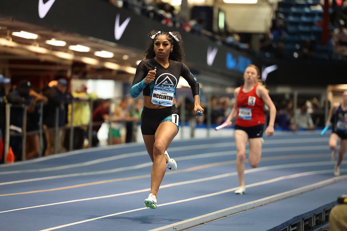 Female sprinter in a black and blue uniform races on a track, baton in hand, during a relay event. Spectators watch in the background.