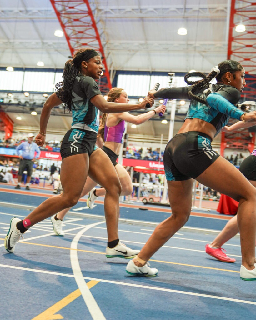 Athletes in a relay race exchange a baton in a sports arena, highlighting teamwork and speed during a competitive event.
