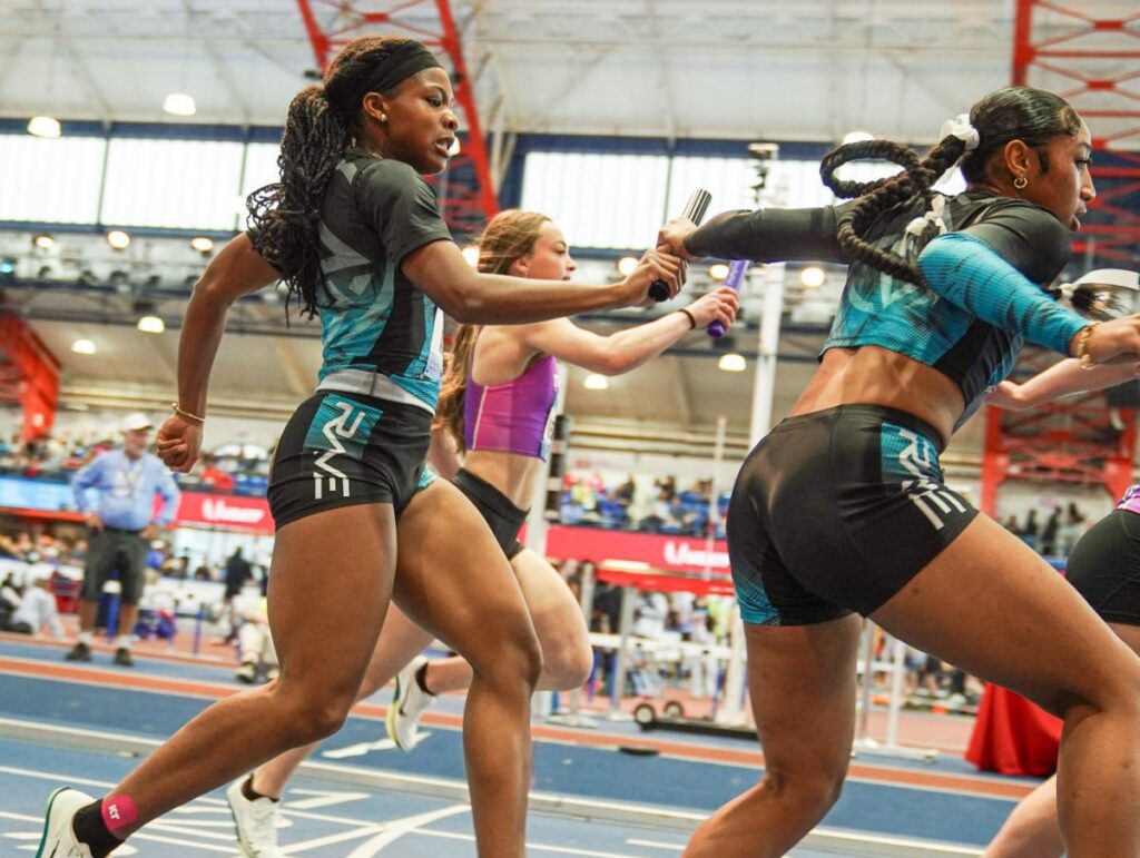 Two female sprinters in motion during a relay race, exchanging a baton in a sports arena filled with spectators.
