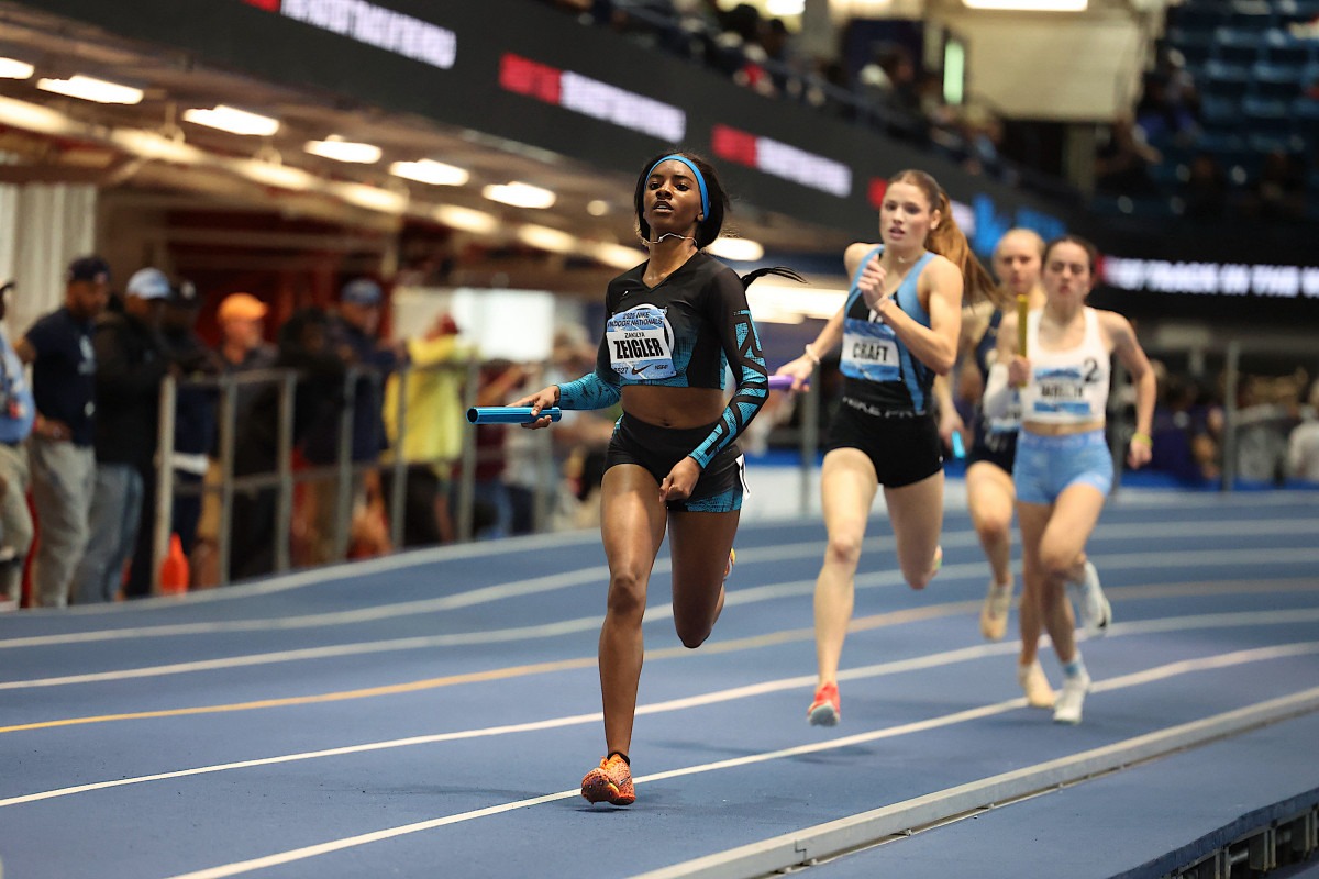 A sprinter in a black and blue uniform is leading a relay race, passing the baton. Two competitors in the background are pursuing closely.