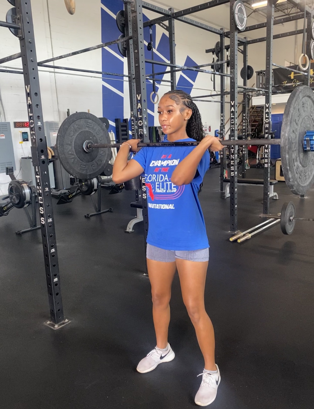 Athlete performing a clean and jerk with a barbell in a gym. Dressed in a blue t-shirt and shorts, she showcases strength training technique.