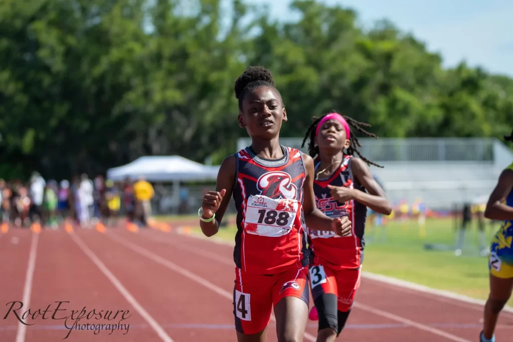 Young athletes compete in a track race under bright sunlight. A girl in red and black gear leads, showing determination.