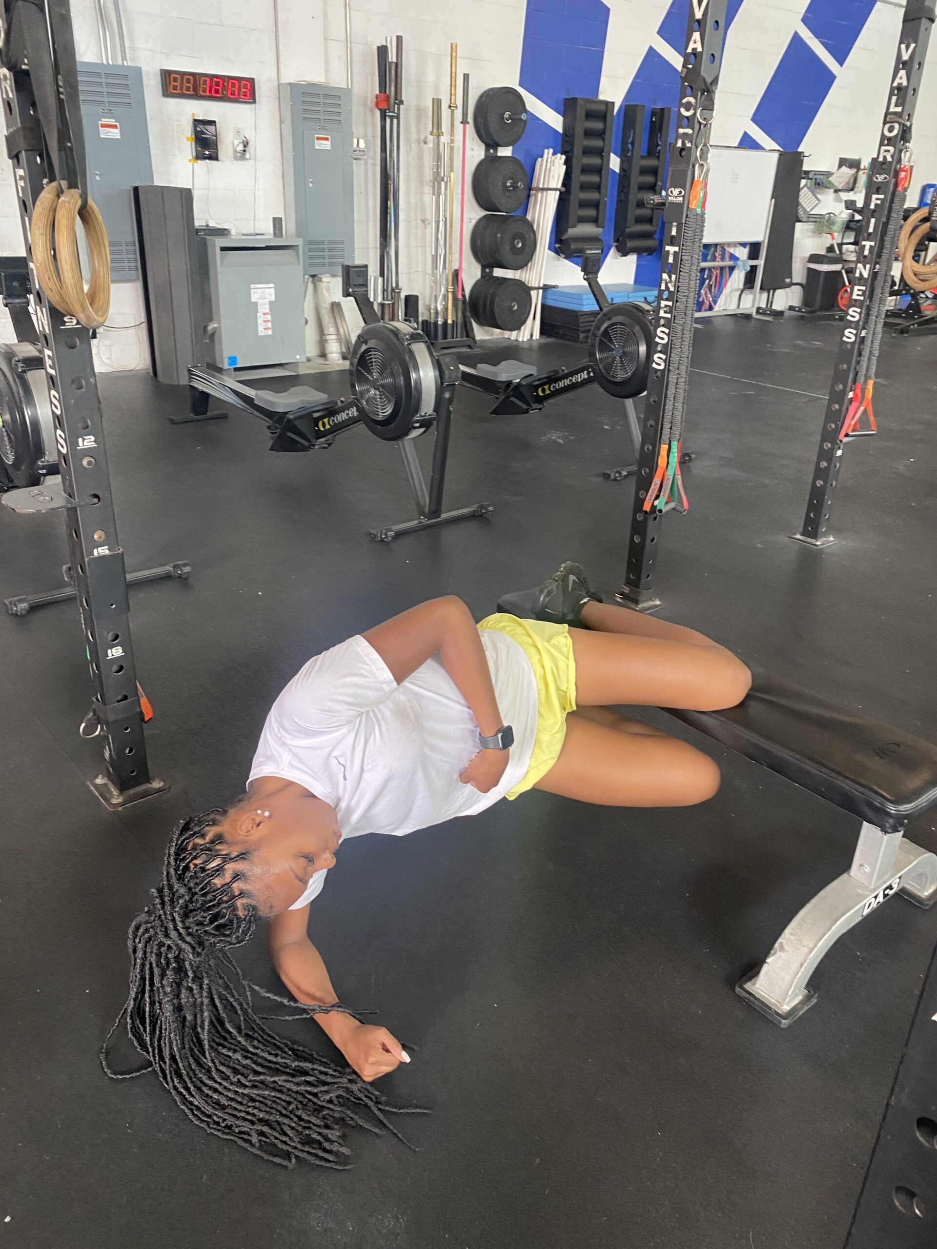 A person with long braided hair lies on a bench in a gym, appearing to rest after a workout. Fitness equipment is visible in the background.