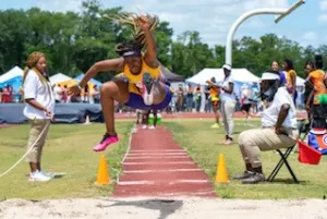 Athlete in mid-air during a long jump, wearing a purple and yellow uniform. Spectators watch from the sidelines at a track and field event.
