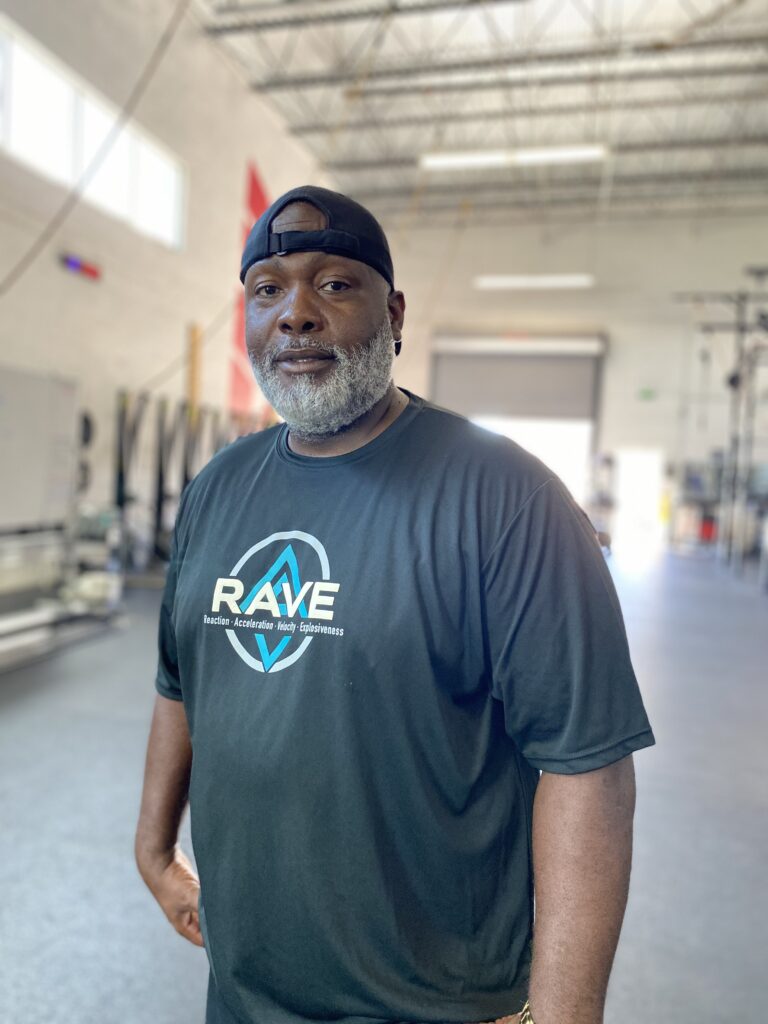 A man with a beard wearing a black RAVE T-shirt and a backward cap stands confidently in a bright, spacious gym, conveying motivation and strength.